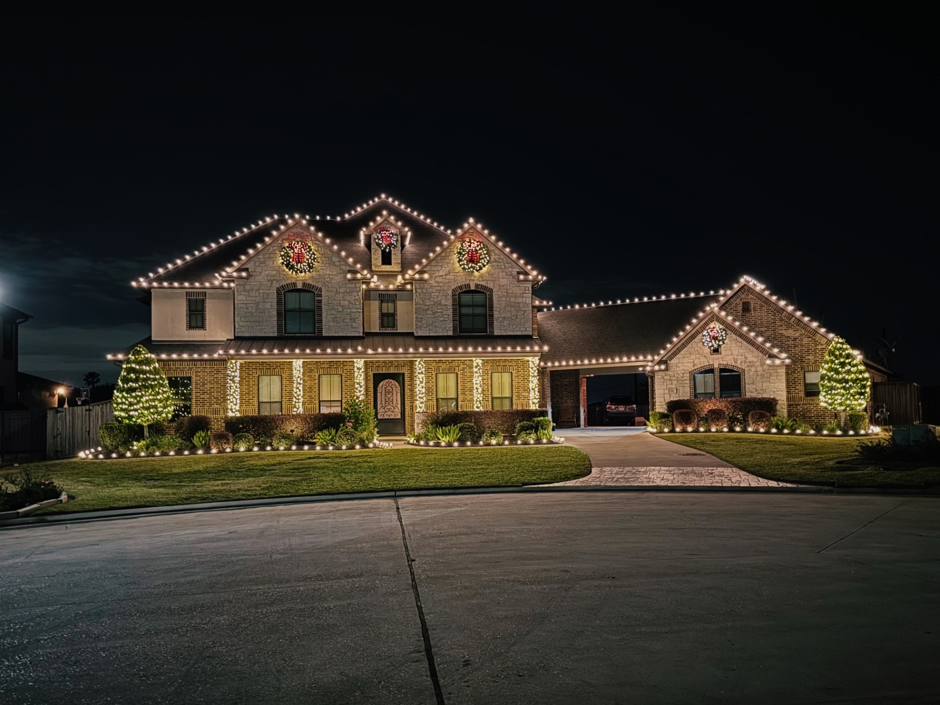 Classic hallmark house with warm white roofline lights, wreaths, and lit trees